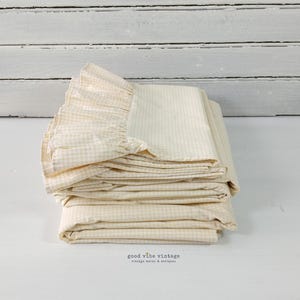May include: A stack of folded cream-colored bedding with a ruffled edge. The fabric has a subtle checkered pattern. The bedding is neatly arranged on a white surface, with a white wooden background.