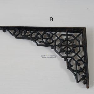 May include: Black, ornate metal shelf bracket with a decorative, openwork design. The bracket features scrolling details and a geometric center. The letter "B" is visible in the upper center. The words "good vibe vintage vintage wares & antiques" are also visible.