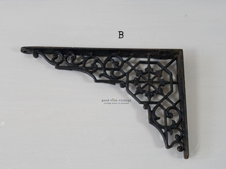May include: Black, ornate metal shelf bracket with a decorative, openwork design. The bracket features scrolling details and a geometric center. The letter "B" is visible in the upper center. The words "good vibe vintage vintage wares & antiques" are also visible.