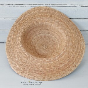 May include: A natural-toned straw hat with a wide brim and a shallow crown. The hat is made of woven straw and has a circular design. The brim is slightly curved, and the hat appears to be unworn. The background is a light-colored wooden surface.