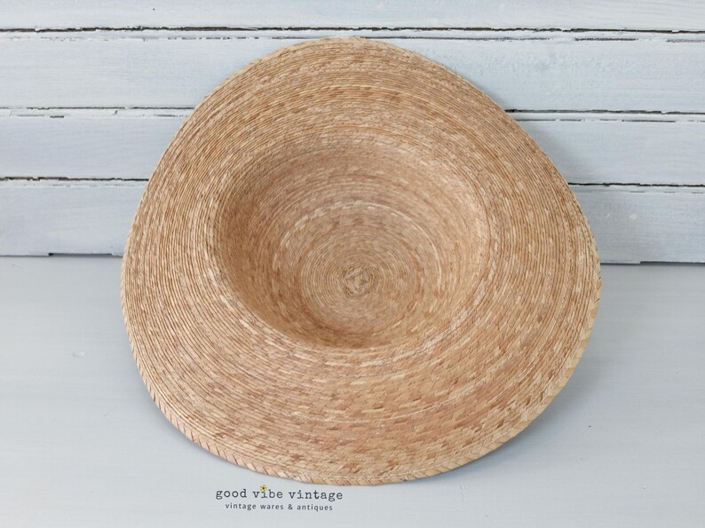 May include: A natural-toned straw hat with a wide brim and a shallow crown. The hat is made of woven straw and has a circular design. The brim is slightly curved, and the hat appears to be unworn. The background is a light-colored wooden surface.