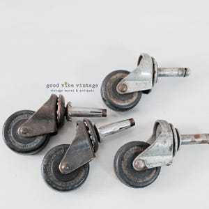 May include: Four vintage metal furniture casters with dark grey rubber wheels and silver-toned mounting stems. Each caster features a swivel bracket and visible wear consistent with age. The text "good vibe vintage" is printed in the background.