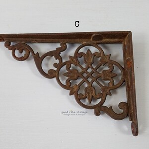 May include: A rusty brown cast iron shelf bracket with an ornate, symmetrical floral design. The bracket is L-shaped, with a decorative scroll on the end. The text "C" is visible on the top. The bracket is from Good Vibe Vintage.