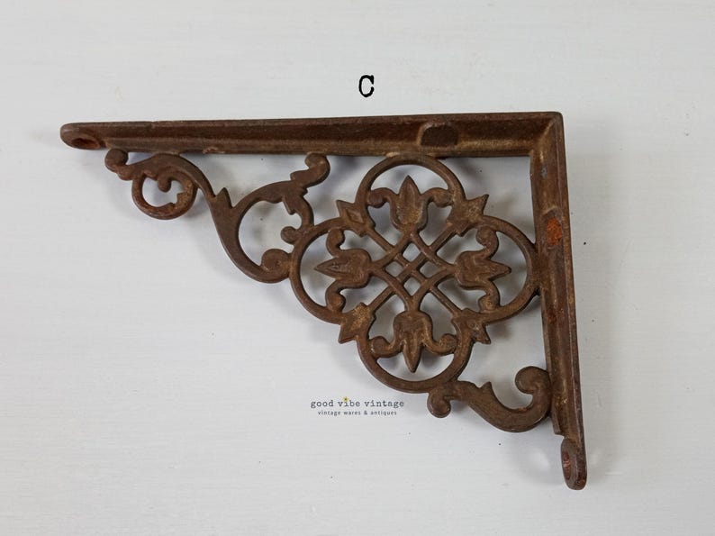 May include: A rusty brown cast iron shelf bracket with an ornate, symmetrical floral design. The bracket is L-shaped, with a decorative scroll on the end. The text "C" is visible on the top. The bracket is from Good Vibe Vintage.