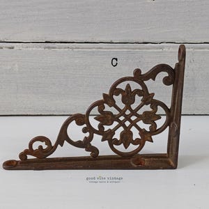 May include: A decorative, cast iron corner bracket with an ornate, openwork design. The bracket is a warm, rusty brown color and features a floral and scroll pattern. The text "C" is visible on the bracket. The words "good vibe vintage" are also visible.