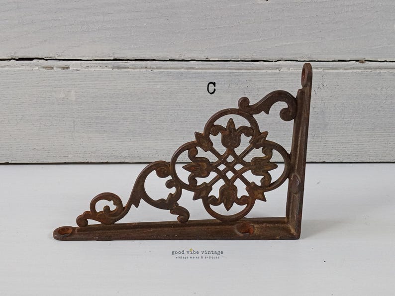 May include: A decorative, cast iron corner bracket with an ornate, openwork design. The bracket is a warm, rusty brown color and features a floral and scroll pattern. The text "C" is visible on the bracket. The words "good vibe vintage" are also visible.