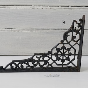 May include: Black, ornate cast iron shelf bracket with decorative scrollwork and a geometric center design. The bracket is in an L-shape, with pre-drilled holes for mounting. The text "B" is visible on the bracket.