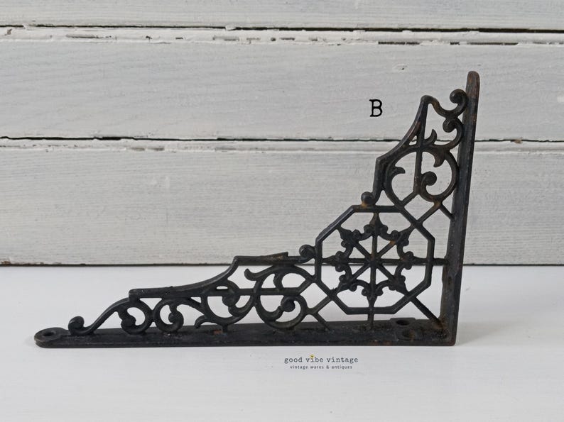 May include: Black, ornate cast iron shelf bracket with decorative scrollwork and a geometric center design. The bracket is in an L-shape, with pre-drilled holes for mounting. The text "B" is visible on the bracket.