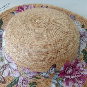 May include: A wide-brimmed sun hat with a natural woven texture. The hat is adorned with a floral and butterfly print in shades of pink, purple, and white. The hat's crown is a lighter shade of tan.