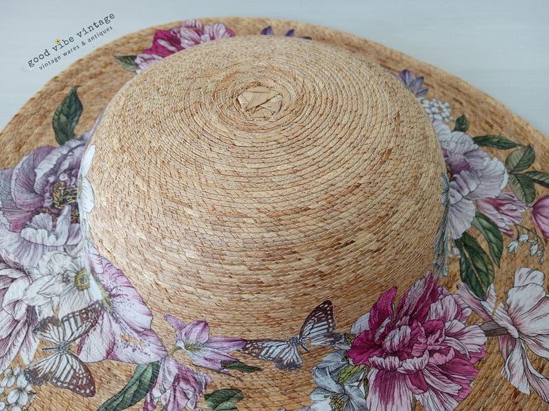 May include: A wide-brimmed sun hat with a natural woven texture. The hat is adorned with a floral and butterfly print in shades of pink, purple, and white. The hat's crown is a lighter shade of tan.