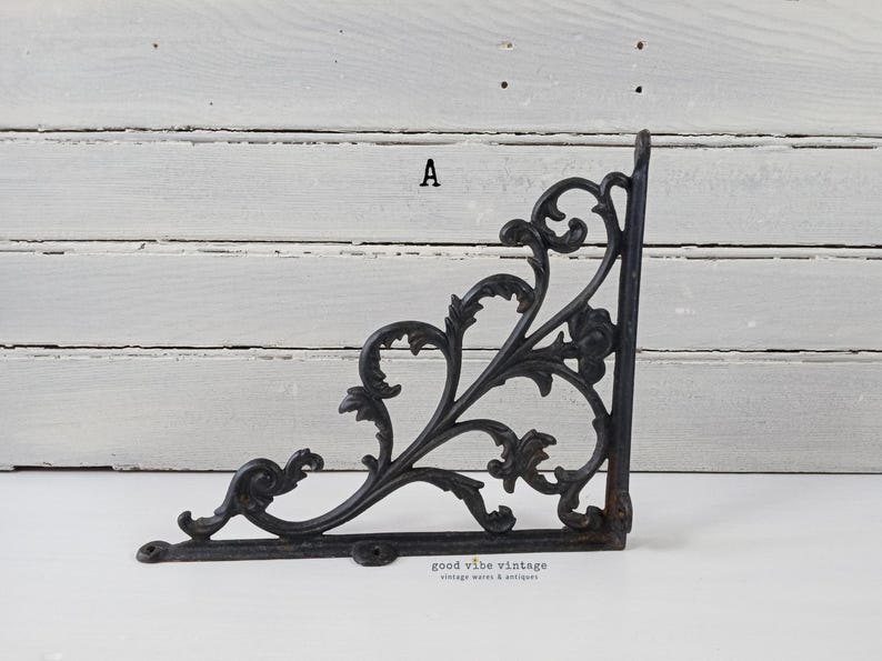 May include: Black, ornate cast iron shelf bracket with a decorative floral and scroll design. The bracket is in the shape of an L, with mounting holes for easy installation. The bracket is a vintage item.