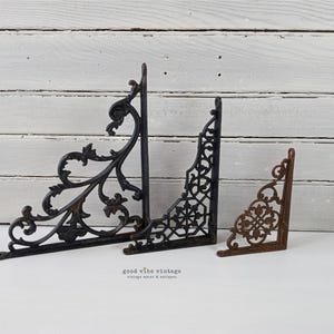 May include: Three decorative metal shelf brackets in varying sizes. The largest is black with an ornate, scrolling design. The middle bracket is black with a geometric pattern. The smallest is a rusty brown color with a floral design.
