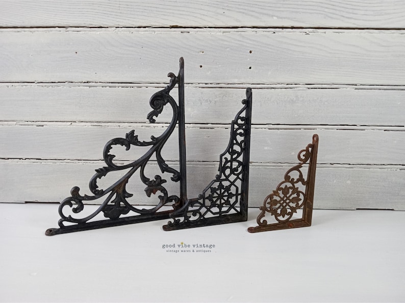 May include: Three decorative metal shelf brackets in varying sizes. The largest is black with an ornate, scrolling design. The middle bracket is black with a geometric pattern. The smallest is a rusty brown color with a floral design.