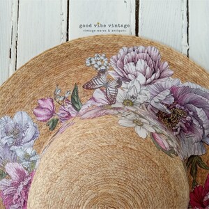 May include: A wide-brimmed sun hat with a natural woven texture, adorned with a floral design featuring purple, pink, and white flowers, and butterflies. The hat has a curved brim and is set against a white wooden background. The text "good vibe vintage" is visible.