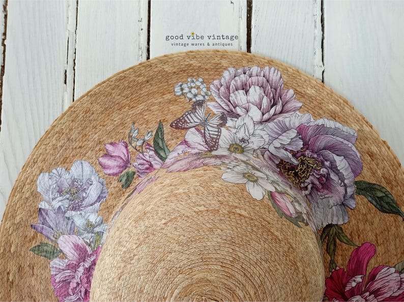 May include: A wide-brimmed sun hat with a natural woven texture, adorned with a floral design featuring purple, pink, and white flowers, and butterflies. The hat has a curved brim and is set against a white wooden background. The text "good vibe vintage" is visible.