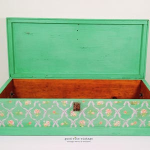 May include: Open, vintage wooden trunk painted in a bright green color. The interior is natural wood. The front of the trunk features a floral pattern with pink roses and white bows. The trunk is open, revealing the interior.