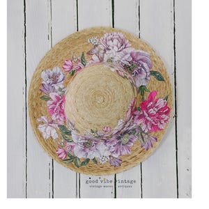 May include: A straw sun hat with a wide brim, adorned with a floral design of pink and purple peonies. The hat is set against a white wooden background. The text "good vibe vintage" is visible.