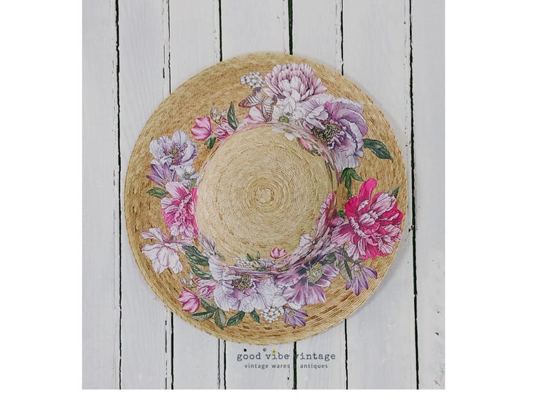 May include: A straw sun hat with a wide brim, adorned with a floral design of pink and purple peonies. The hat is set against a white wooden background. The text "good vibe vintage" is visible.