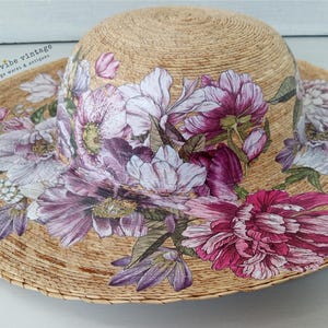 May include: A wide-brimmed sun hat made of woven straw, adorned with a floral design. The flowers are in shades of pink, purple, and white, with green leaves. The hat has a natural straw color and is suitable for sun protection.