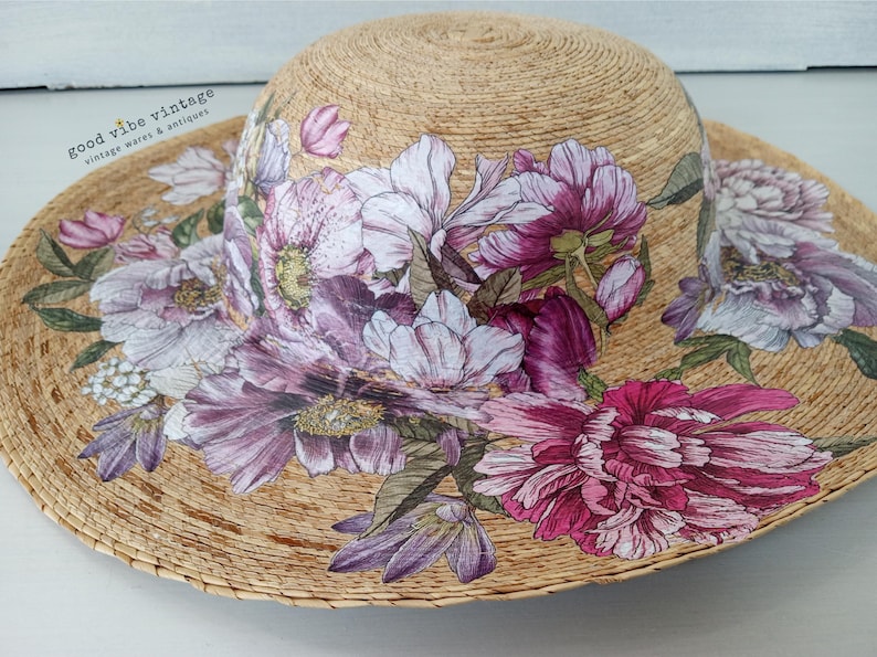 May include: A wide-brimmed sun hat made of woven straw, adorned with a floral design. The flowers are in shades of pink, purple, and white, with green leaves. The hat has a natural straw color and is suitable for sun protection.