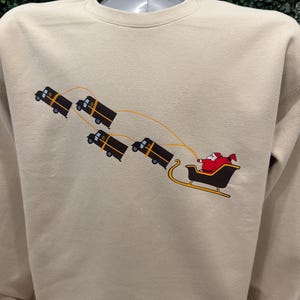 May include: A beige sweatshirt featuring a graphic of Santa Claus riding in a sleigh pulled by four delivery trucks. The trucks are black with yellow accents and the sleigh is black with red accents.