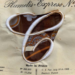 Op de afbeelding: Een geborduurde witte letter "C" met een crèmekleurige rand, op een beige papieren label. Het label leest "Plumetis-Express N" en "Made in France". De achtergrond heeft een bruin en oranje bloemenpatroon.