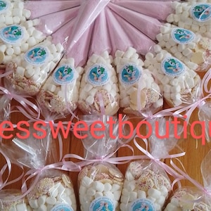 May include: An arrangement of cellophane bags filled with white marshmallows and cookies, tied with pink ribbons. Each bag has a circular sticker with a cartoon image and text. The bags are arranged around pink cones.