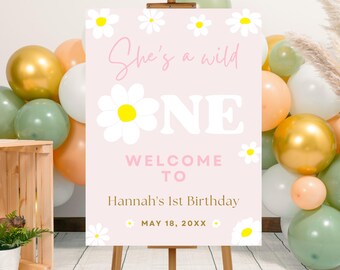 Op de afbeelding: Een roze welkomstbord op een ezel met de tekst "She's a wild ONE" en "Welcome to Hannah's 1st Birthday". Het bord is versierd met madeliefjes en omringd door gouden, witte, perzik- en groene ballonnen.
