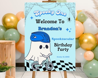 May include: A light blue sign on an easel with the text "Spooky One Welcome To Brandon's Spooktacular Birthday Party". A cartoon ghost wearing a blue helmet rides a bicycle. Balloons in gold, white, orange, and green surround the sign.