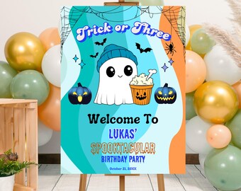 May include: A Halloween-themed party sign on an easel. The sign reads "Trick or Three" and "Welcome to Lukas' Spooktacular Birthday Party." The sign features a cartoon ghost, pumpkins, bats, and popcorn.