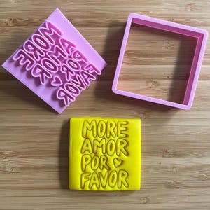 May include: A light purple square cookie stamp with the embossed text MORE AMOR POR FAVOR and a heart symbol, a matching light purple square cookie cutter frame, and a yellow square cookie with the same embossed text and heart symbol.