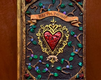 May include: A rectangular book with a dark brown textured cover featuring a central red heart emblem with gold filigree, surrounded by sculpted vines, green leaves, and small purple flowers. A banner reads "JACK & JOHNNY" and "EST 2025".