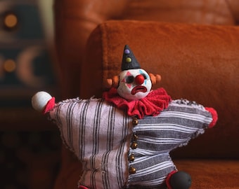May include: A star-shaped clown doll featuring a white face with red and blue painted details, a red nose, and a black pointed hat. The body is made of black and white striped fabric with a red ruffled collar, small metal bells, and round black feet.