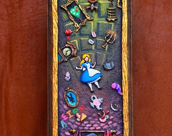 May include: A rectangular wall plaque with a gold-painted wooden frame featuring a relief scene of a girl in a blue dress surrounded by floating objects including a clock, playing cards, a teapot, a mirror, a candle holder, and a small door.
