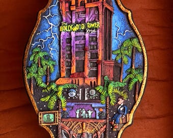 May include: A decorative wall plaque featuring a relief sculpture of The Hollywood Tower Hotel. The scene includes a dark building, lightning bolts, palm trees, and various small figures. The text reads The Hollywood Tower Hotel in yellow.
