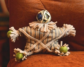 May include: A stuffed fabric doll shaped like a mummy with a round head wrapped in beige strips. The body features a blue and white striped pattern with beige fabric strips crisscrossed over the torso. It has frayed fabric limbs and green beads.