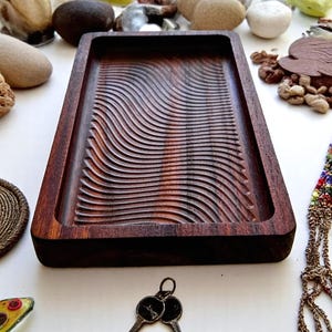 May include: A rectangular, dark brown wooden tray with a carved, wave-like pattern. The tray is made of a rich, dark wood and has a smooth, polished finish. The tray is surrounded by small stones and decorative items.