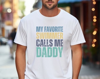 May include: White t-shirt with the text "My Favorite Swimmer Calls Me Daddy" in a distressed font with blue, yellow, and green colors.