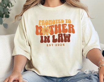 May include: A white t-shirt with the text "Promoted to Mother in Law Est 2024" in a retro font. The text is in brown and orange with a small flower graphic.