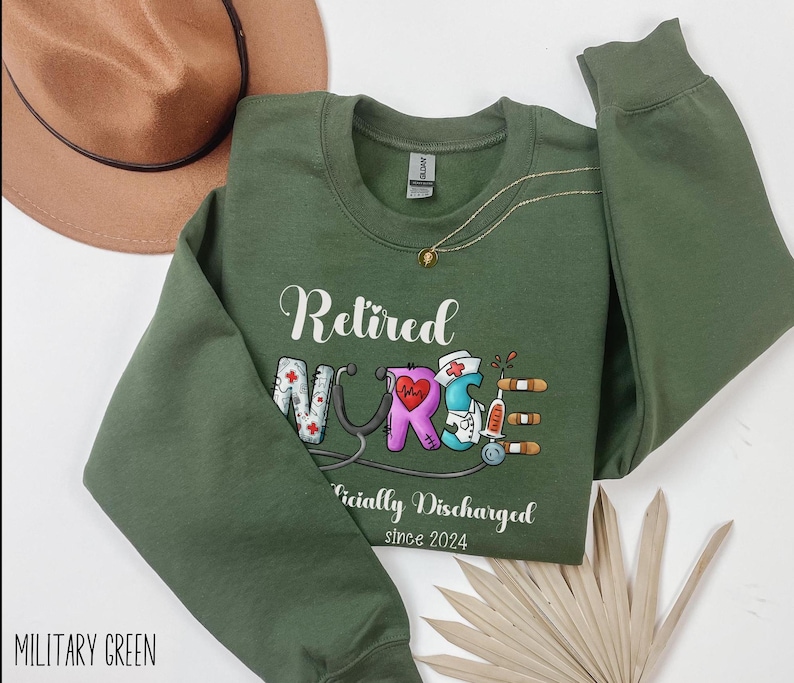 May include: A military green sweatshirt with the text "Retired Nurse Officially Discharged since 2024" and a colorful illustration of a nurse's uniform.