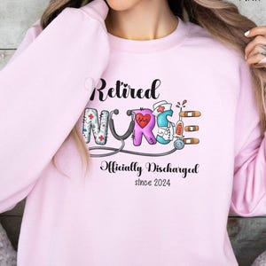 May include: Pink sweatshirt with the text "Retired Nurse" in black lettering. The word "Nurse" is illustrated with colorful medical symbols. The text "Officially Discharged since 2024" is below the word "Nurse".