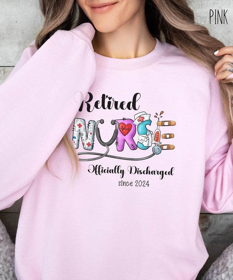 May include: Pink sweatshirt with the text "Retired Nurse" in black lettering. The word "Nurse" is illustrated with colorful medical symbols. The text "Officially Discharged since 2024" is below the word "Nurse".
