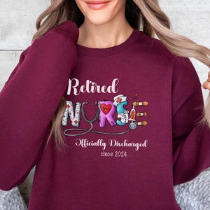May include: Maroon sweatshirt with the text "Retired Nurse" in white lettering. The word "Nurse" is decorated with medical symbols and the text "Officially Discharged since 2024" is below.