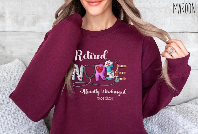 May include: Maroon sweatshirt with the text "Retired Nurse" in white lettering. The word "Nurse" is decorated with medical symbols and the text "Officially Discharged since 2024" is below.