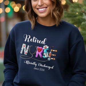 May include: Navy blue sweatshirt with the text "Retired Nurse Officially Discharged since 2024" and a graphic of a stethoscope and medical tools forming the word "Nurse".
