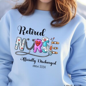 May include: Light blue sweatshirt with the text "Retired Nurse Officially Discharged since 2024" and a colorful graphic of a stethoscope and medical symbols.