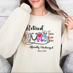 May include: A cream-colored sweatshirt with the text "Retired Nurse Officially Discharged since 2024" and a colorful illustration of the word "Nurse" with medical symbols.