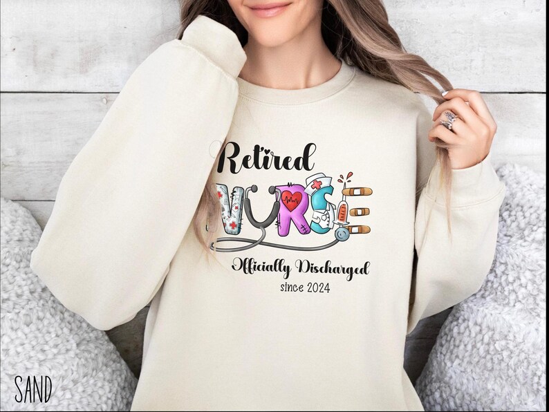 May include: A cream-colored sweatshirt with the text "Retired Nurse Officially Discharged since 2024" and a colorful illustration of the word "Nurse" with medical symbols.