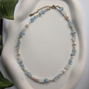 Könnte beinhalten: Eine zarte Halskette mit einer Kette aus hellblauen Aquamarin-Chips, weißen Perlen und kleinen goldfarbenen Perlen. Die Halskette wird auf einem weißen, organisch geformten Tablett präsentiert, das ihr natürliches und elegantes Design hervorhebt.