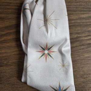 May include: Folded off-white fabric with a repeating starburst design in gold, pink, blue, and green. The fabric has a subtle sheen. The item is likely a scarf or headband.